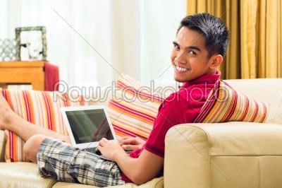 Asian man sitting on couch surfing the internet 