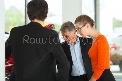 Mature man with young couple and auto in car dealership are looking beneath a hood