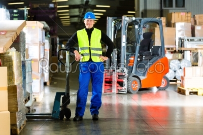 Warehouseman in protective vest holds a mover, standing beside packages and boxes at warehouse of freight forwarding company- a forklift is in Background