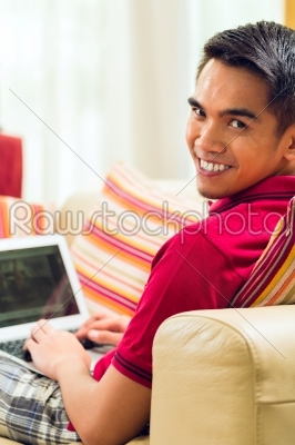Asian man sitting on couch surfing the internet 