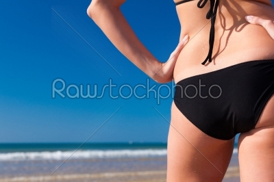 Attractive Woman in bikini standing in the sun on beach, only part of torso to be seen, a lot of copyspace in the blue sky