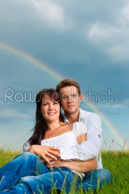 Happy couple sitting on a meadow under rainbow