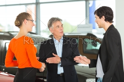 Mature man handshaking with young couple and auto in car dealership