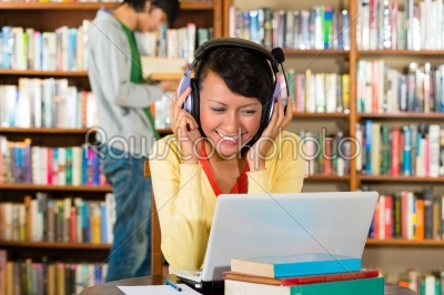 Student - Young woman in library with laptop and headphones learning, a male student standing in the Background reads a book