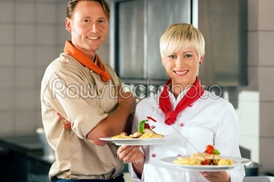 Two chefs in teamwork - man and woman - in a restaurant or hotel kitchen cooking delicious food, both are presenting the finished dishes