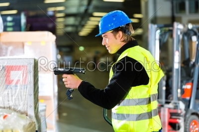 Warehouseman in protective vest using a scanner, standing beside packages and boxes at warehouse of freight forwarding company- a forklift is in Background