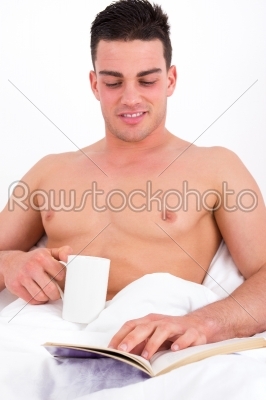 handsome man holding cup of coffee and reading in bed